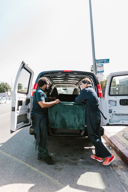 Two movers from Man with Van Putney are engaged in a home relocation, standing behind an open van with its back doors wide open on a paved street. They are lifting and loading a large, rectangular pie