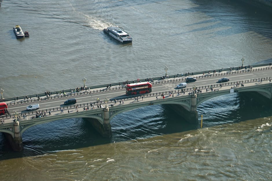 A wide view of the River Thames in Putney, taken during daytime under a partly cloudy sky. The river is dotted with various boats and small vessels, with some moored near the riverbank while others ar