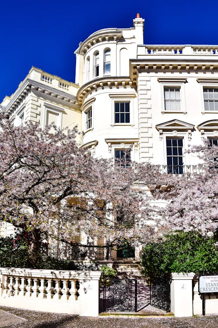 The image shows a residential house with a light grey exterior and white window frames, partly obscured by a large cherry blossom tree in full bloom with pink flowers and some green leaves. The house features a front door with the number 6, and is accessed via a small brick and concrete staircase flanked by black metal railings. In front of the house, there is a neatly trimmed, dense green hedge that runs along a low brick wall. The scene is set outdoors during daytime, with natural light illuminating the scene. This setting is relevant to house removals, as it depicts the typical environment where a home relocation or furniture transport might take place, reflecting the importance of careful handling of belongings in front of residential properties. Man with Van Putney’s removals service might operate in this area, supporting clients with packing, loading, and transportation of items during a move or spring clearout.